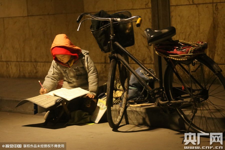 鄭州中原路京廣鐵路大橋下涵洞的馬路牙子邊，一位戴著紅色辣椒帽的小男孩趴在一個小馬扎上，上面鋪著一張車墊，在微弱的燈光下寫作業(yè)