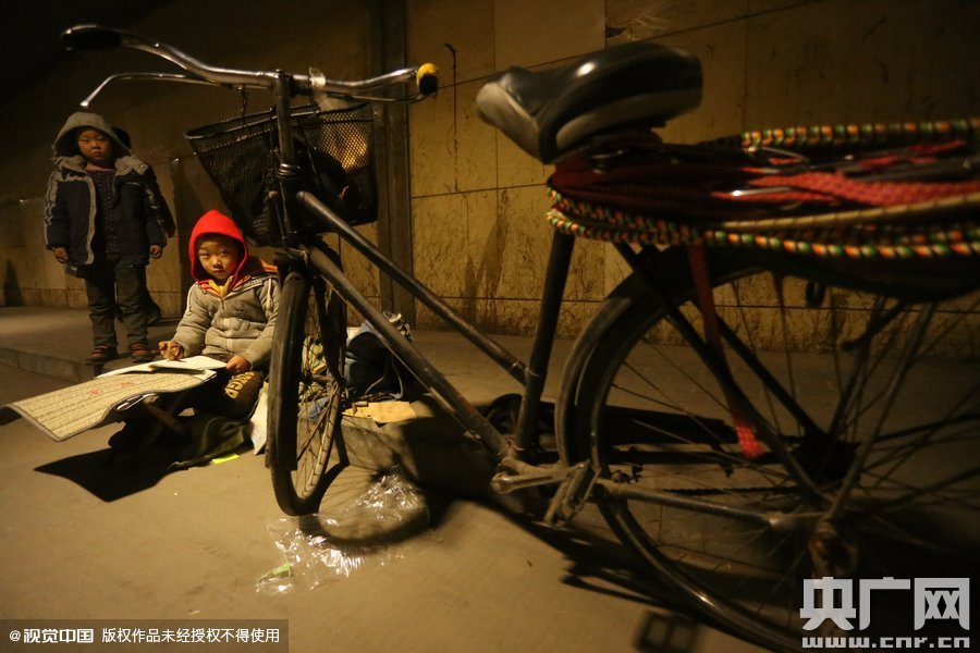 2015年11月8日晚，鄭州，7歲男孩涵洞橋下陪父母練攤，路邊燈下坐小馬扎上寫作業(yè)。
